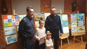 Policjanci z nagrodzonymi dziewczynkami na tle prac konkursowych.