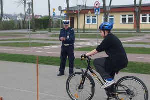 Dziecko na rowerze, w tle policjant weryfikujący poprawność przejazdu w ramach turnieju „JEDNOŚLADEM BEZPIECZNIE DO CELU”