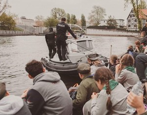 Policjanci na policyjnej łodzi motorowej płyną rzeką Brdą w rejonie Opery Nowa. Na brzegu siedzą harcerze i ich obserwują.