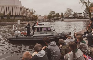 Rzeka Brda przy Operze Nowa. Policjanci na policyjnej łodzi motorowej płyną rzeką Brdą. Na brzegu siedzą harcerze i ich obserwują.
