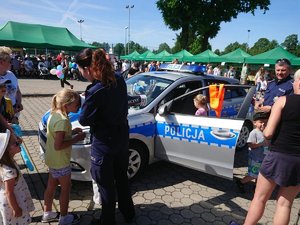 Policyjny radiowóz stojący na tle namiotów. Stojąca przy nim policjantka pokazuje dzieciom i dorosłym &quot;magiczną&quot; tubę.