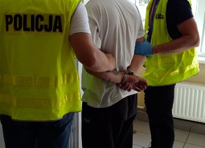 Ujęcie kajdanek na rękach zatrzymanego, którego prowadzą policjanci.