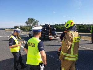 Policjanci i strażak ustalają okoliczności zdarzenia, w tle laweta z wrakiem samochodu