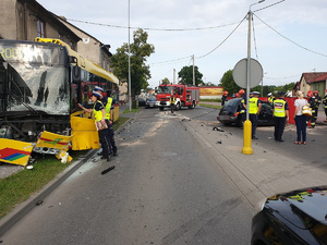 Rozbity autobus i osobowy chrysler, policjanci i strażacy pracują na miejscu wypadku na miejscu.