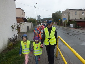 Policjantka idzie z uczniami ubranymi w odblaski po chodniku