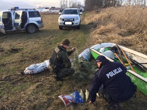 Policjant i strażnik rybacki kucają w pobliżu łodzi klusownika, obok złowione szczupaki, a w tle policyjny radiowóz i auto sprawcy