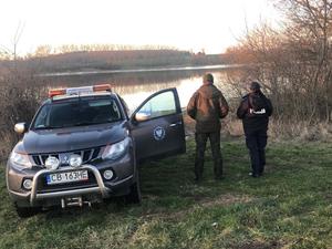 Policjantka i strażnik łowiecki stoją obok radiowozu nad jeziorem