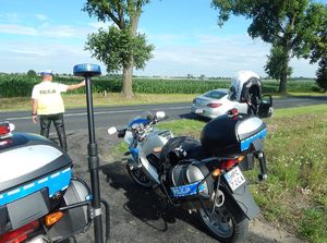 Policjant kontroluje miernikiem prędkość nadjeżdżającego kierowcy, przed nim dwa oznakowane motocykle