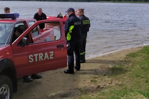 strażacy i policjant stoją przy samochodzie straży pożarnej przed brzegiem jeziora