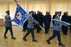 poczet sztandarowy wychodzi z auli. Trzech policjantów idzie jeden za drugim. W tle stoją nowoprzyjęci policjanci