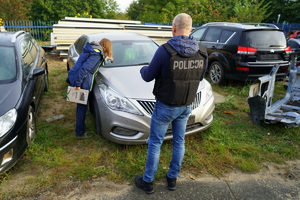 policjant stoi z przodu szarego pojazdu i wykonuje oględziny pisząc na protokole. Policjantka zagląda na podszybie pojazdu