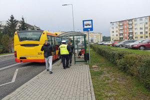 Policjant oraz inne osoby idą w stronę autobusu, który stoi na przystanku autobusowym.