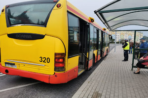 Policjant i inne osoby stoją na przystanku autobusowym. Widoczny stojący autobus miejski.