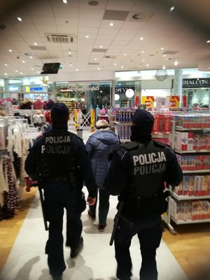 Kobieta w asyście policjantów idzie aleją sklepową