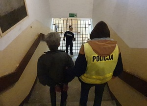 Policjant prowadzi zatrzymanego po schodach. Przed nimi widoczne kraty oraz inny policjant.