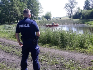 Policjant stoi nad małym jeziorkiem, a na wodzie pływa strażak łodzią