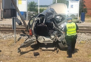 Policjant stoi przy rozbitym aucie na przejeździe kolejowym.