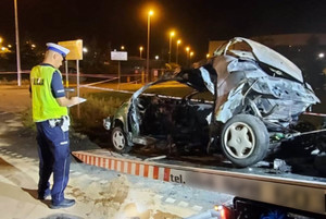 Policjant stoi nad spalonym wrakiem samochodu który jest wciągany na lawetę