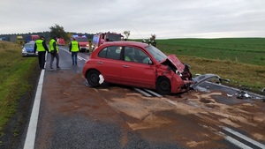 Rozbity nissan obok idą policjanci, a w tle stoją radiowozy i wozy strażackie