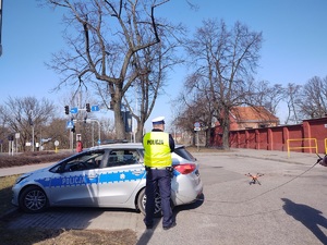 Policjant stoi przy radiowozie, a obok na ziemi stoi dron