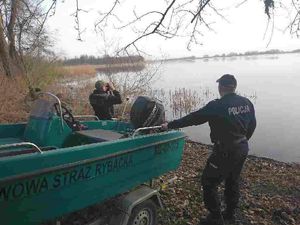 Pracownik straży rybackiej i policjant stoją na brzegu akwenu. Strażnik rybacki obserwuje zbiornik wodny przy pomocy lornetki. Obok nich stoi łódź.