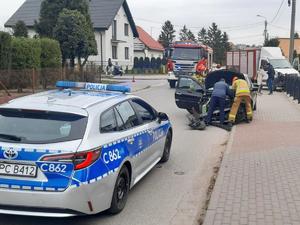 Na pierwszym planie zdjęcia stoi radiowóz oznakowany, przed nim stoi samochód ze zniszczonym przodem. Przy samochodzie stoją trzy osoby: dwóch strażaków i osoba cywilna.  Za samochodem stoi radiowóz straży pożarnej oraz po jego lewej stronie auto dostawcze, które wjechało w ogrodzenie posesji.