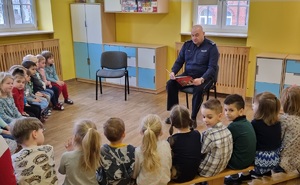 Siedzący na krześle policjant, który czyta książkę siedzącym na ławkach dzieciom.