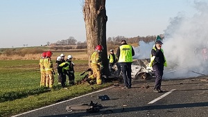 Widok na rozbity i spalony samochód osobowy oraz pracujące na miejscu służby ratunkowe