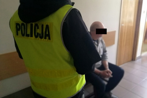 Mężczyzna siedzi na ławce. Obok niego stoi policjant.