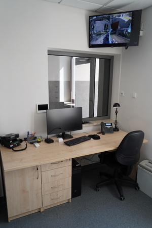 ujęcie na pokój dyżurki z biurkiem, na którym stoi monitor, klawiatura, telefon, radiostacja, a na ścianie jest przeszklone okno do przyjmowania petentów