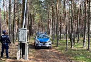 Policjant stoi obok słupa, z którego zostały wycięte kable.  W pobliżu zaparkowany jest radiowóz