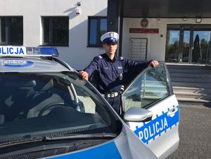 Policjant stoi przy radiowozie. W tle budynek jednostki.