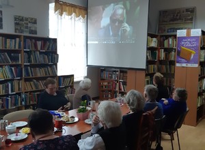 Policjant stoi przy ekranie projekcyjnym na przeciwko seniorom siedzącym w ławkach