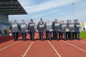 policjanci maszerują w tyralierze w dwuszeregu na płycie stadionu ubrani w sprzęt ochrony osobistej. Z przodu idą policjanci z tarczami