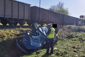 Policjant RD robi oględziny rozbitego samochodu, za nimi stoi pociąg towarowy