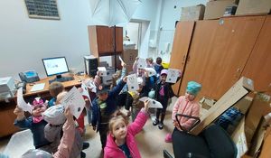 Dzieci stoją w grupie w pomieszczeniu do daktyloskopi i trzymają w rękach kartki z odciskami palców