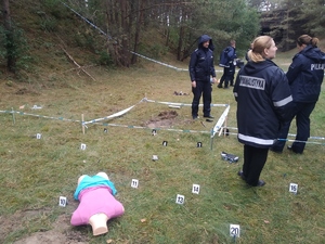 Miejsce wybuchu. Na pierwszym planie leży ludzki korpus, tabliczki znamionowe, rozciągnięta taśmą, stoi kilku policjantów. W pobliżu szczątków technik kryminalistyki.