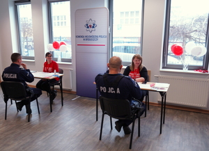 Przy dwóch ławkach siedzą dwie kobiety i dwóch policjantów. Pomiędzy nimi stoi baner z napisem Komenda Wojewódzka Policji w Bydgoszczy