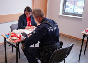 Przy ławce siedzi kobieta po cywilnemu, na przeciwko niej siedzi policjant. Na ławce leżą czerwone smycze, koperty i formularze.