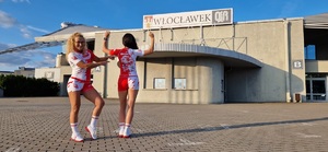 Dwie kobiety (jedna z nich to policjantka z Włocławka) w strojach o barwach biało-czerwonych stoją na tle zabudowań z napisem Włocławek