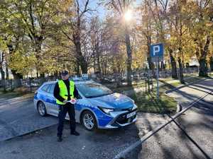 Policjant stoi przy radiowozie zaparkowanym przy cmentarzu