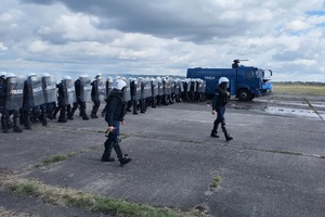 policjanci ćwiczą na płycie lotniska. Funkcjonariusze ustawieni są w szeregu. Pomiędzy nimi jedzie policyjny pojazd z wodą