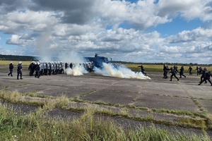 policjanci ćwiczą na płycie lotniska. Funkcjonariusze ustawieni są w szeregu. Pomiędzy nimi jedzie policyjny pojazd z wodą