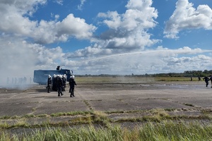 policjanci ćwiczą na płycie lotniska. Funkcjonariusze ustawieni są w szeregu. Pomiędzy nimi jedzie policyjny pojazd, który leje strumieniem wody w kierunku osób