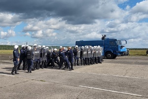 policjanci ćwiczą na płycie lotniska. Funkcjonariusze ustawieni są w szeregu. Pomiędzy nimi jedzie policyjny pojazd, który leje strumieniem wody w kierunku osób
