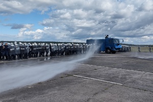 policjanci ćwiczą na płycie lotniska. Funkcjonariusze ustawieni są w szeregu. Pomiędzy nimi jedzie policyjny pojazd, który leje strumieniem wody w kierunku osób