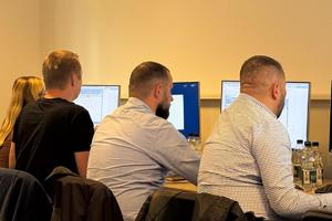 nieumundurowani policjanci siedzą przed komputerami i słuchają prowadzącego szkolenie