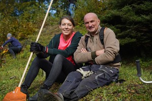 Uśmiechnięta kobieta i mężczyzna siedzą na trawie w czasie przerwy, trzymając narzędzia ogrodnicze. Za nimi inni uczestnicy akcji nadal porządkują teren na wzniesieniu.