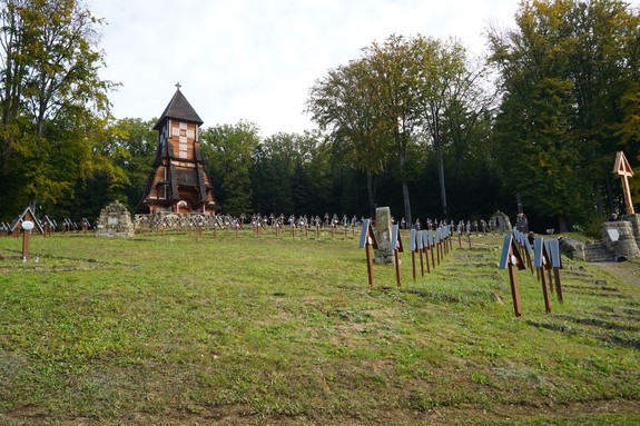 Na pierwszym planie widać wysoką drewnianą wieżę-kaplicę cmentarza, a przed nią rzędy zadbanych, jednakowych krzyży. Cały teren jest wykoszony i uporządkowany.