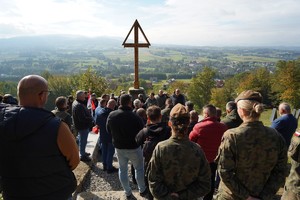 Grupa uczestników akcji stoi przy dużym drewnianym krzyżu i słucha przemówienia osoby prowadzącej. Za nimi rozpościera się rozległy widok na pola, domy i wzgórza.